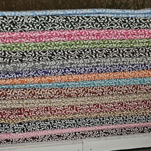 Puede incluir: Una pila de piezas de tela dobladas, cada una con un patrón constante de hojas blancas en espiral sobre un fondo sólido. Los colores incluyen azul claro, negro, rosa, verde, marrón oscuro, gris, morado, naranja, turquesa, beige, rojo y rosa claro.