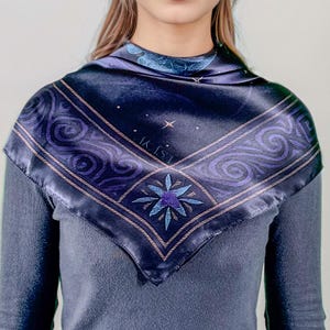 May include: A dark blue square scarf with a celestial design. The scarf features a central starburst motif in shades of blue and purple, surrounded by swirling patterns and the text "IK VI". The scarf is worn over a gray long-sleeved shirt.