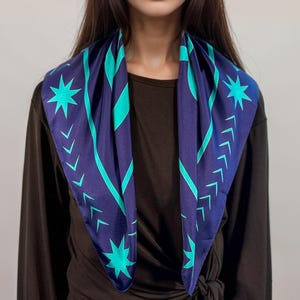 May include: A square scarf in navy blue with a teal star and arrow pattern. The scarf is draped around a person's neck, showcasing the geometric design. The scarf is made of a soft material.