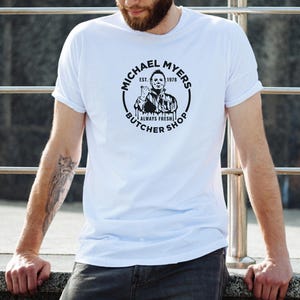 May include: White t-shirt with a black and white graphic of Michael Myers from the Halloween movies. The graphic says "Michael Myers Butcher Shop Est. 1978 Always Fresh".