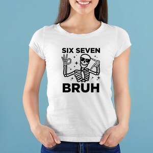 May include: White t-shirt with a black skeleton graphic. The skeleton is wearing sunglasses and making hand gestures. The text on the shirt reads "SIX SEVEN BRUH".