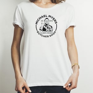 May include: White t-shirt with a black and white graphic of Michael Myers, the character from the Halloween movies. The graphic includes the text "Michael Myers Est. 1978 Always Fresh Butcher Shop".