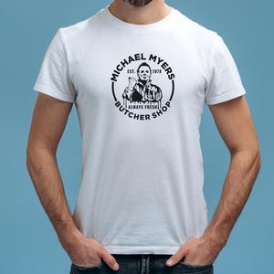 May include: White t-shirt with a black and white graphic of Michael Myers from the movie Halloween. The graphic is a circle with the text "Michael Myers Butcher Shop Est. 1978 Always Fresh" around the image of Michael Myers.