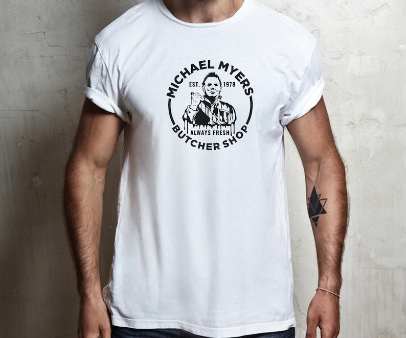May include: White t-shirt with a black and white graphic of Michael Myers from the Halloween franchise. The graphic says "Michael Myers Butcher Shop Est. 1978 Always Fresh".