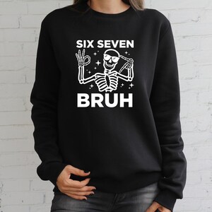 May include: Black sweatshirt with a white graphic of a skeleton wearing sunglasses and making hand gestures. The text "SIX SEVEN BRUH" is above and below the skeleton illustration. The sweatshirt is a casual style.