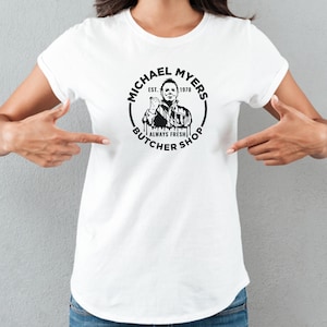 May include: White t-shirt with a black and white graphic of Michael Myers from the movie Halloween. The graphic says "Michael Myers Butcher Shop Est. 1978 Always Fresh".