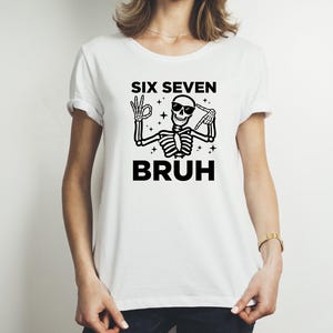 May include: White t-shirt featuring a black skeleton graphic wearing sunglasses and making hand gestures. The text "SIX SEVEN BRUH" is above and below the skeleton illustration. The shirt has short sleeves and a crew neck.