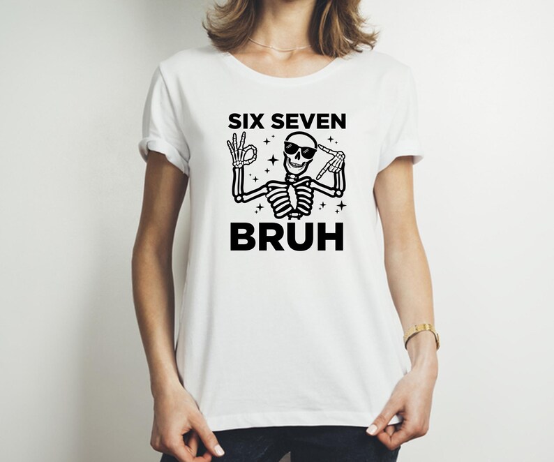 May include: White t-shirt featuring a black skeleton graphic wearing sunglasses and making hand gestures. The text "SIX SEVEN BRUH" is above and below the skeleton illustration. The shirt has short sleeves and a crew neck.