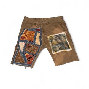 May include: Brown denim shorts with patchwork details. The shorts feature various fabric patches in shades of brown, red, and blue. One patch has a leaf design. The shorts have a distressed look.