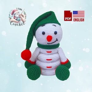 Peut inclure: Un jouet de bonhomme de neige en crochet avec un chapeau vert, des mitaines rouges et un corps blanc. Le bonhomme de neige est assis avec un sourire sur son visage.