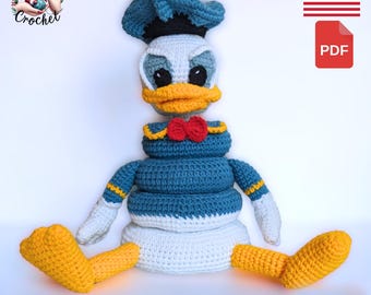 Op de afbeelding: Een handgemaakte gehaakte Donald Duck pop in blauw, wit, geel en rood. De pop heeft een blauwe matrozenhoed, een gele snavel en een rode vlinderdas. De pop zit met uitgestrekte benen. De tekst "I can Crochet" is inbegrepen.