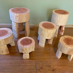 May include: Six wooden stools with a natural wood finish and a reddish-brown top. The stools are all different sizes and have three legs each.