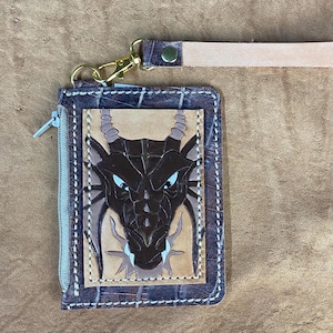 May include: Brown leather wallet with a dragon design. The wallet has a zipper closure and a gold-colored clasp with a matching leather strap. The dragon is brown with blue eyes and is centered on a tan background.