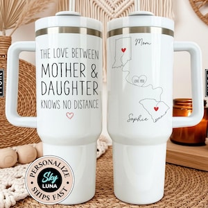 May include: White travel mug with a handle, featuring text that reads "THE LOVE BETWEEN MOTHER & DAUGHTER KNOWS NO DISTANCE." The back of the mug has a map design with hearts and the words "Mom" and "Sophie."