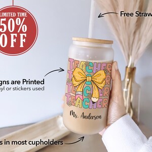 May include: A frosted glass tumbler with a colorful "Teacher" design featuring a yellow bow and a pencil. The tumbler has a bamboo lid and a straw. The text "Ms. Anderson" is printed on the bottom of the tumbler.