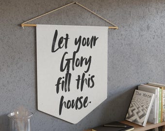 Puede incluir: Una pancarta blanca con el texto "Let your Glory fill this house" en escritura negra cuelga de una pared. La pancarta está suspendida por una varilla de madera y cuerda. Una estantería de madera debajo contiene libros y un cuaderno.