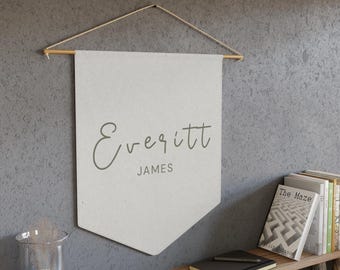May include: A cream-colored banner with the name "Everitt" and the word "JAMES" printed in a script font. The banner is suspended from a wooden dowel with a natural cord. The banner hangs on a gray wall.