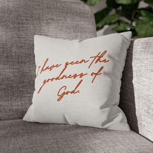 May include: A square, light gray pillow with the phrase "I have seen the goodness of God" in a rust-colored script. The pillow is placed on a gray upholstered chair.