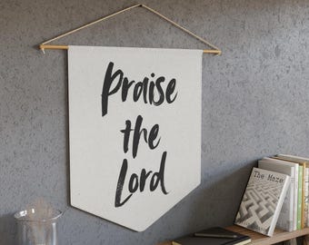 Puede incluir: Una pancarta de lona blanca con el texto "Praise the Lord" escrito en caligrafía negra. La pancarta está colgada de una varilla de madera con cuerda.