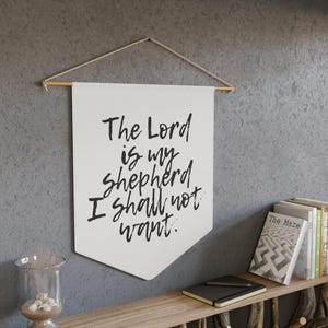 May include: A white banner with the text "The Lord is my shepherd I shall not want" in black script. The banner is suspended from a wooden dowel with a natural cord. The banner hangs against a gray wall.