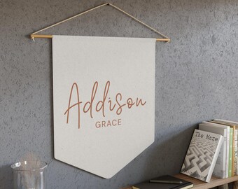 May include: Cream-colored wall banner with the name "Addison" in a copper-colored script font, with "Grace" below. The banner hangs from a wooden dowel and twine. A wooden shelf below holds books, a notebook, and a glass vase.