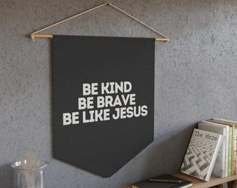 Puede incluir: Bandera negra con el texto blanco "BE KIND BE BRAVE BE LIKE JESUS". La bandera cuelga de una barra de madera con una cuerda. La bandera cuelga de una pared gris.