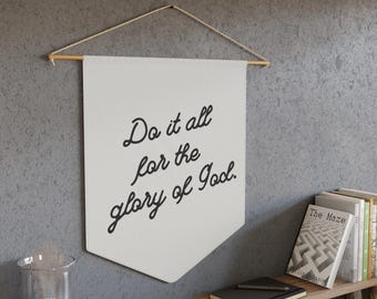 Puede incluir: Bandera de lona blanca con el texto "Do it all for the glory of God." colgada de una pared con una varilla de madera en la parte superior. La bandera tiene forma de banderín con la parte inferior puntiaguda.