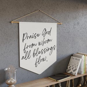 Puede incluir: Un cartel blanco con el texto "Praise God from whom all blessings flow" en escritura negra. El cartel está suspendido de una varilla de madera y cuerda. Una estantería de madera debajo contiene libros, un cuaderno y un portavelas de cristal.