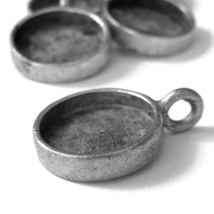 May include: Close-up of several small, silver-colored, oval-shaped bezel settings with a loop for attaching a chain or other findings. The settings have a shallow, concave interior for holding a cabochon or other embellishment.