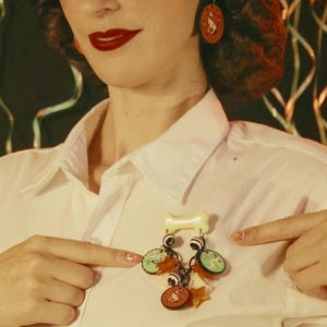 May include: A pair of round, dark brown earrings featuring a white cat illustration, and a brooch consisting of a cream-colored bone-shaped top piece with dangling charms, including small round discs with cat graphics, black and white beads, and stars.