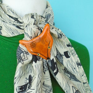 May include: A cream-colored scarf with a black and white pattern is secured with an orange fox-shaped brooch. The brooch has gold-colored eyelets and is positioned at the center of the scarf. The scarf is draped over a green garment.