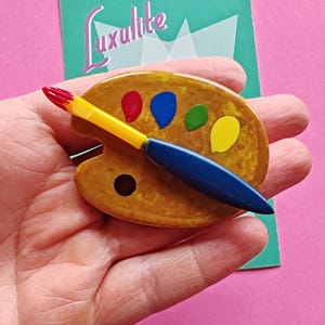May include: A vintage artist's palette brooch with a yellow and brown base, featuring dabs of red, blue, green, and yellow paint. A blue and yellow paintbrush rests on the palette. The word "Luxulite" is on a teal card in the background.