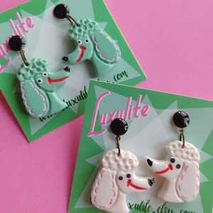May include: Earrings featuring poodle dog designs in mint green and cream colors. Each earring has a black stud and a red smile. The earrings are displayed on green cards with the text "Luxulite.etsy.com".