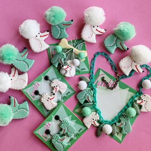 May include: A collection of poodle-themed jewelry and accessories on a pink background. Items include earrings, brooches, and a necklace, featuring white and mint green poodles with fluffy pom-pom accents. The text "Luxulite_etsy.com" is visible on some items.