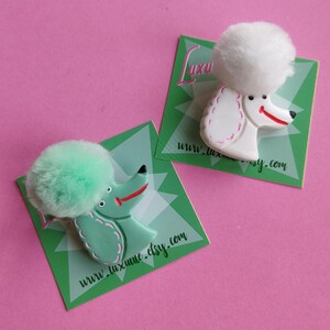 Puede incluir: Dos broches con forma de caniche, uno blanco y otro verde menta, cada uno con una cabeza de pomp&oacute;n esponjosa. Los broches est&aacute;n sobre tarjetas verdes con el texto "www.luxume.etsy.com" y un fondo rosa.