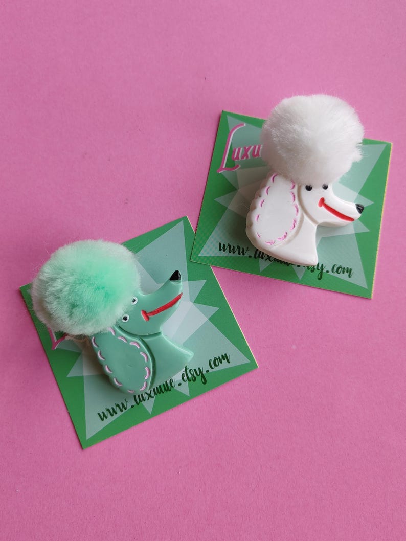 Puede incluir: Dos broches con forma de caniche, uno blanco y otro verde menta, cada uno con una cabeza de pomp&oacute;n esponjosa. Los broches est&aacute;n sobre tarjetas verdes con el texto "www.luxume.etsy.com" y un fondo rosa.