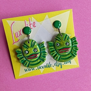Puede incluir: Un par de pendientes de criatura verde con labios rojos y ojos blancos, con un diseño colgante. Los pendientes están sujetos a una tarjeta amarilla con el texto Luxlite y www.luxulite.etsy.com impreso en rosa y verde.