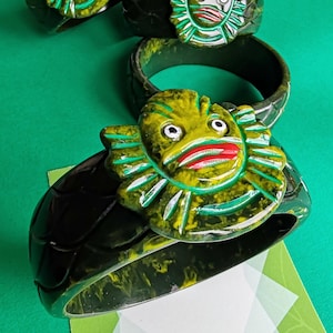 May include: Several dark green resin bracelets with a creature design in yellow, green, and red. The creature has large eyes and a wide mouth. The bracelets are on a green surface.
