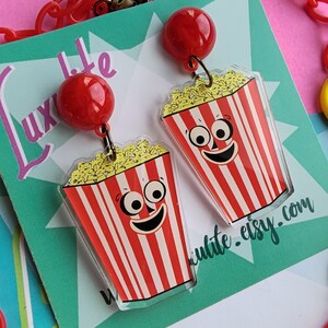 May include: Novelty earrings with a popcorn design. Each earring has a red and white striped popcorn bucket with a smiling face and a red ball stud. The earrings are on a teal background with the text "Luxlite Life" and "luxlite.etsy.com".