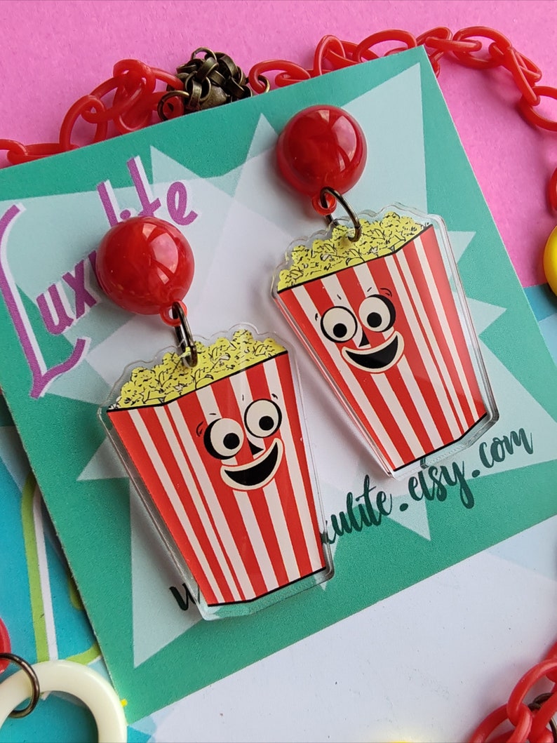 May include: Novelty earrings with a popcorn design. Each earring has a red and white striped popcorn bucket with a smiling face and a red ball stud. The earrings are on a teal background with the text "Luxlite Life" and "luxlite.etsy.com".