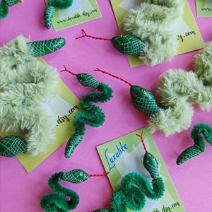 Puede incluir: Varios broches con forma de serpiente que presentan cuerpos verdes difusos, cabezas verdes lisas con patrones de cuadrícula blancos y lenguas de alambre rojo fino. Cada broche está sujeto a una tarjeta verde claro con el texto Luxlite.