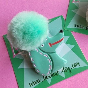 Puede incluir: Broche de caniche verde menta con un pomp&oacute;n esponjoso y detalles rosas. El broche est&aacute; sobre una tarjeta verde con el texto "www.luxume.etsy.com". El fondo es rosa.