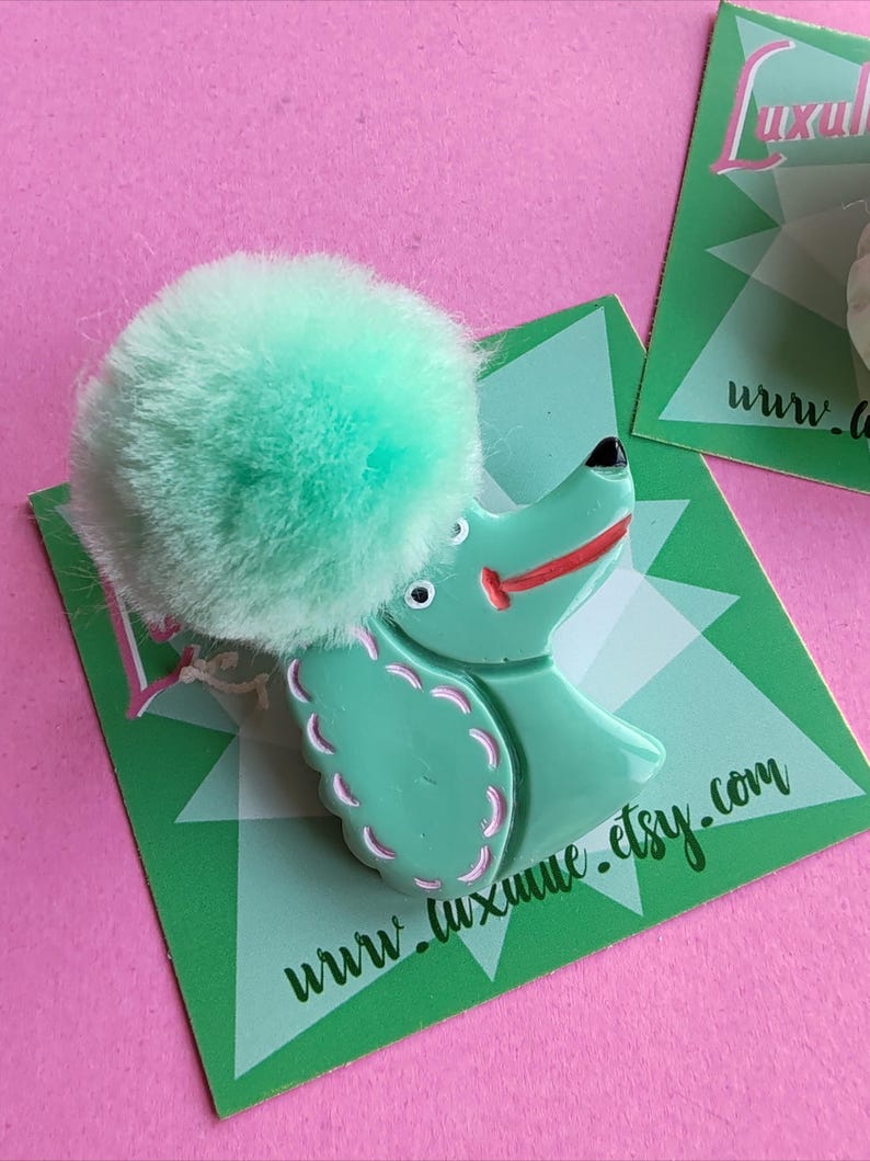 Puede incluir: Broche de caniche verde menta con un pomp&oacute;n esponjoso y detalles rosas. El broche est&aacute; sobre una tarjeta verde con el texto "www.luxume.etsy.com". El fondo es rosa.