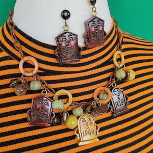 May include: A necklace and matching earring set featuring brown tombstone charms with painted faces, pink cheeks, and crossbones. The necklace includes orange and yellow plastic rings, green pumpkin beads, and a yellow tombstone charm marked 1949.