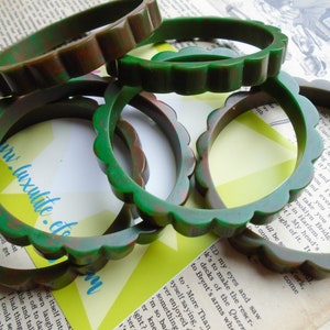 May include: A collection of vintage green and brown scalloped bangles. The bracelets are made of a mottled material, with a wavy edge design. The background includes vintage book pages and a yellow and green geometric shape.