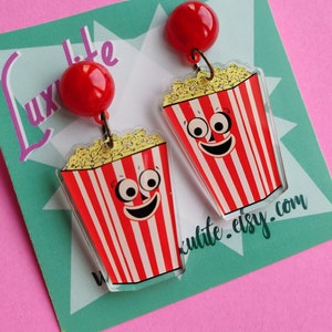 May include: Novelty earrings with a popcorn design. Each earring has a red and white striped popcorn container with a smiling face and a red ball at the top. The earrings are set against a teal and pink background.