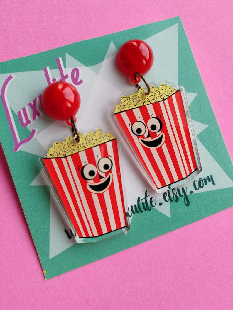 May include: Novelty earrings with a popcorn design. Each earring has a red and white striped popcorn container with a smiling face and a red ball at the top. The earrings are set against a teal and pink background.