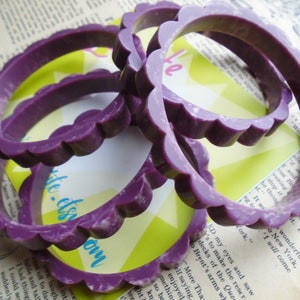May include: Several purple bangles with a scalloped edge design are stacked on top of each other. The bracelets are a solid color with a slightly marbled appearance. The background includes a vintage book page and a yellow and green card.