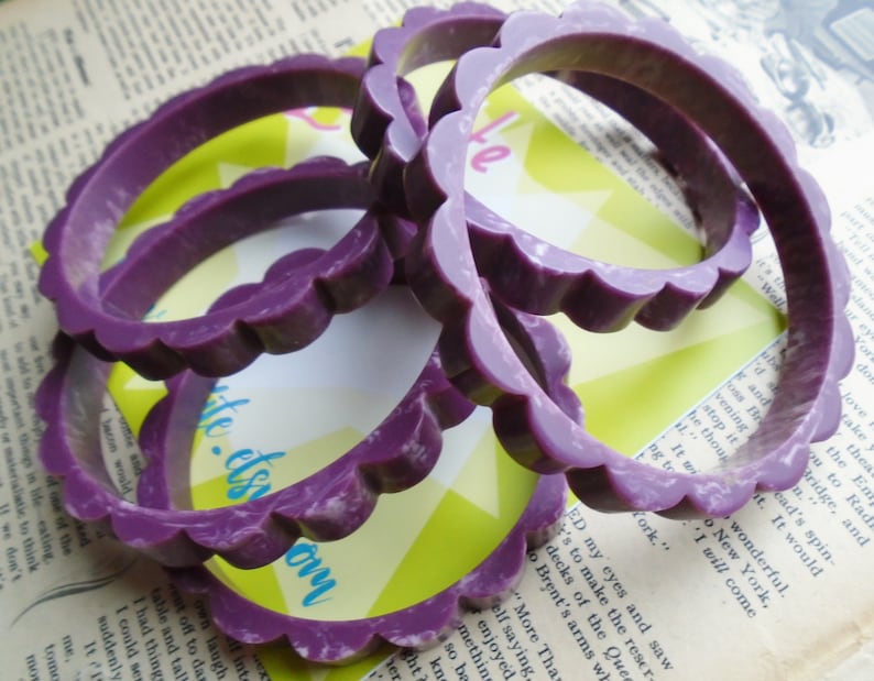 May include: Several purple bangles with a scalloped edge design are stacked on top of each other. The bracelets are a solid color with a slightly marbled appearance. The background includes a vintage book page and a yellow and green card.