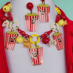 May include: A quirky jewellery set with a necklace and earrings. The necklace and earrings feature popcorn box charms with smiling faces, red and white stripes, and yellow popcorn. The necklace has red chain links, yellow and red beads.